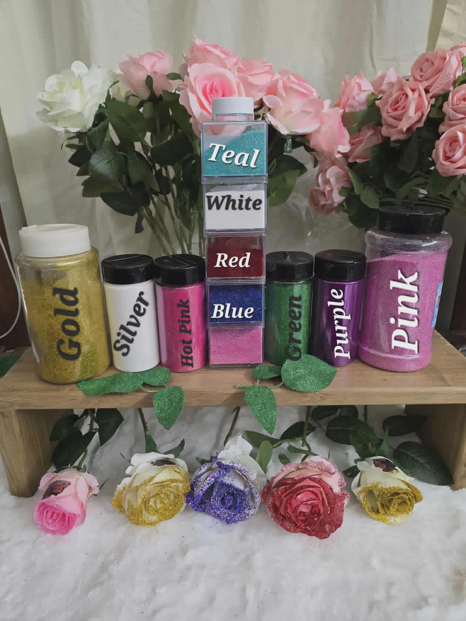 Colorful glitter jars with labels on a wooden stand, surrounded by flowers.