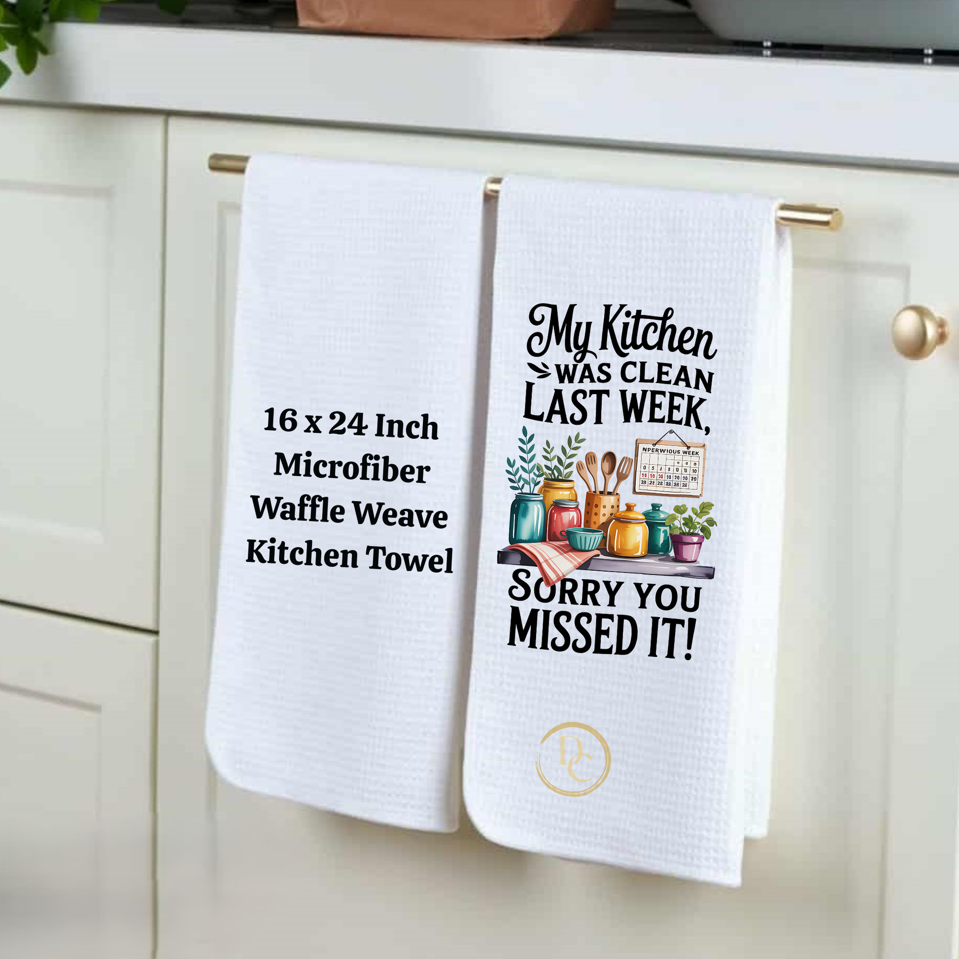 Two white kitchen towels hanging on a cabinet handle, one with text and the other with a graphic design.